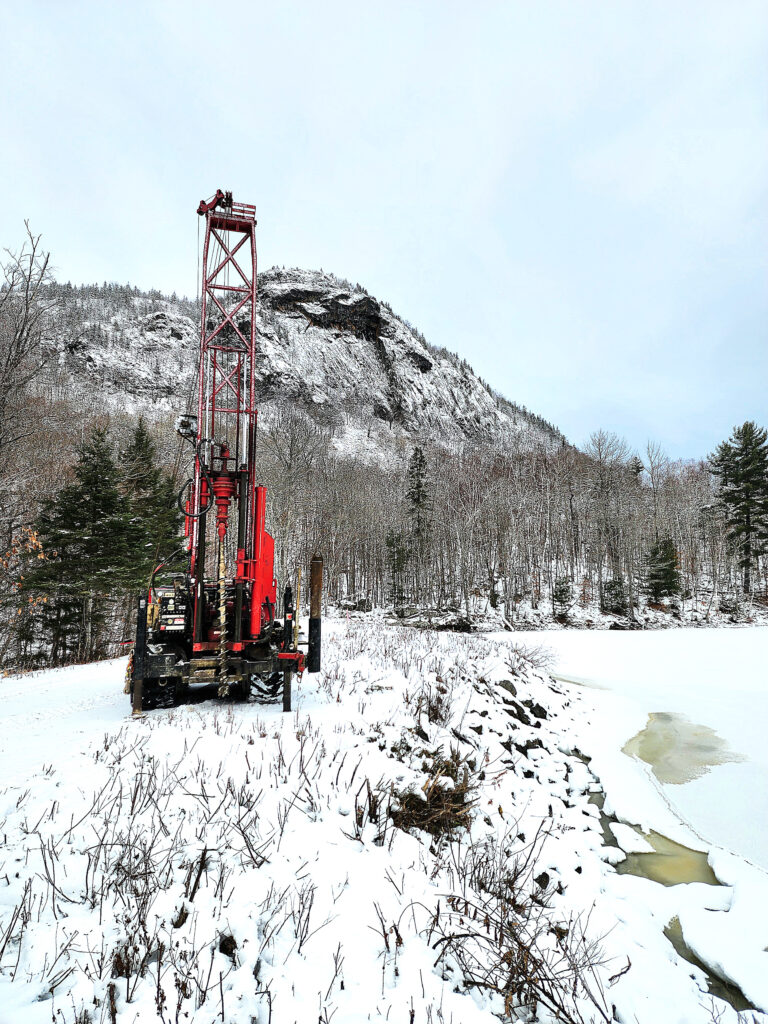 Drill rig in snow with snow covered mountain in background and frozen body of water beside.