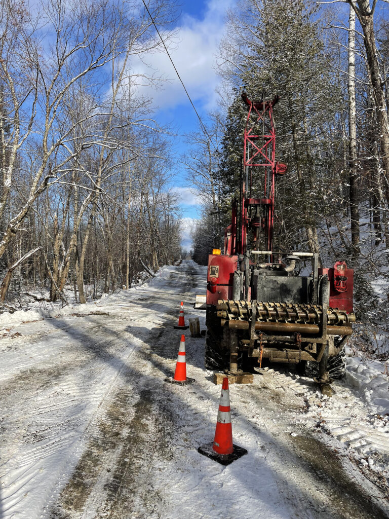 Drill rig parked on side of road in a snow covered forest.