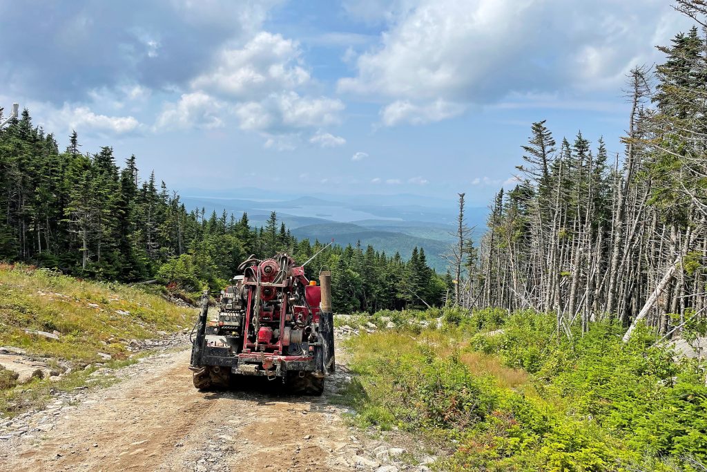 Drilling equipment traveling on dirt road with trees beside and view of mountains, hills and water in the distance
