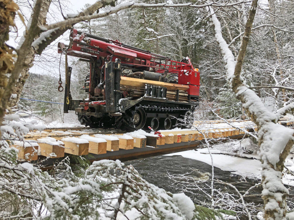 Drill rig on bridge above stream with snow covered trees around