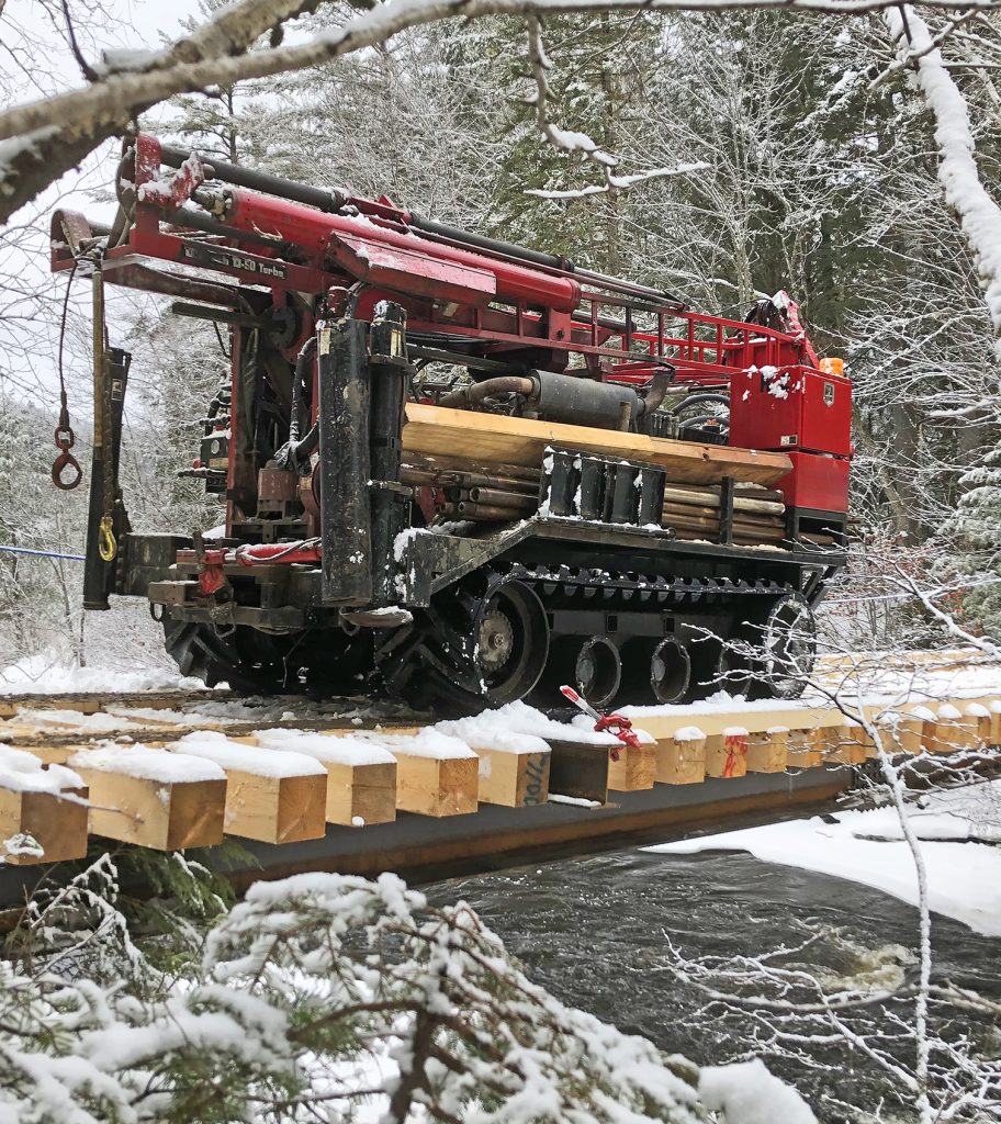 Drill rig on bridge above stream with snow covered trees around