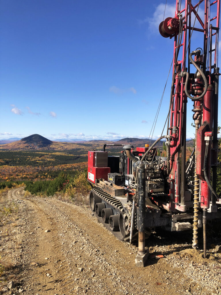Photo of a drill rig on a dirt road with mountains in the distance.