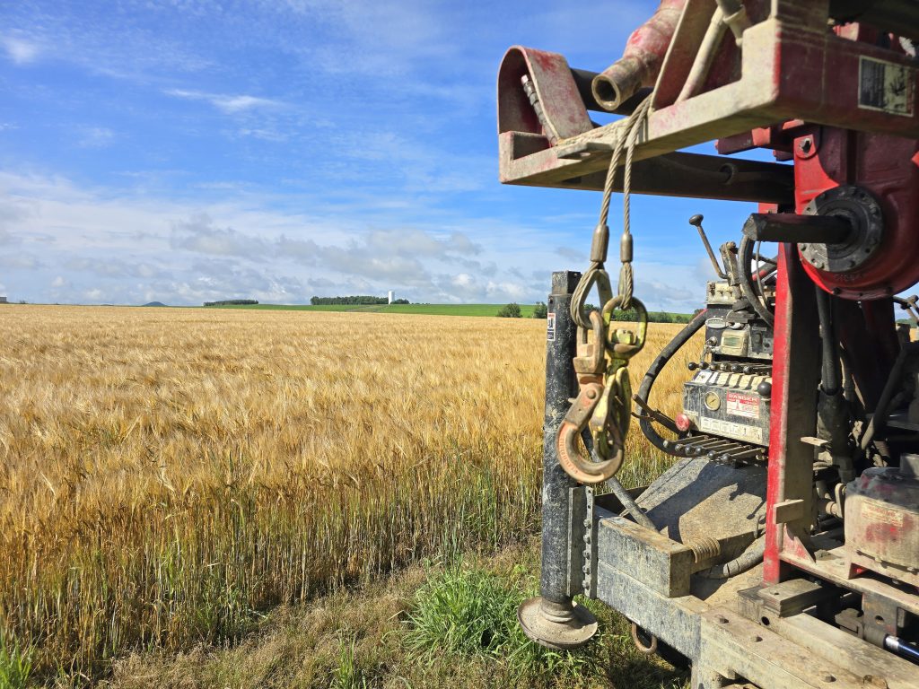 The back of a geotechnical drilling rig with a farm field in the background.