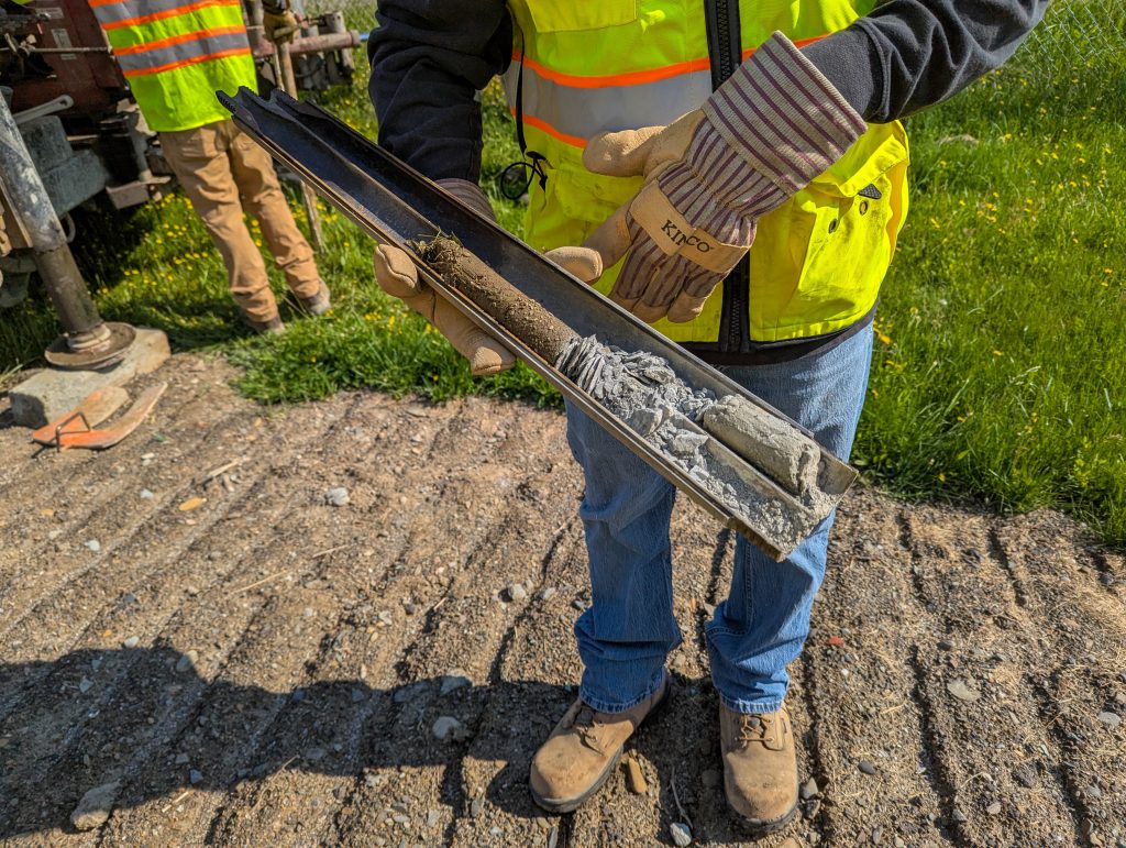 A geotechnical engineer wearing PPE holds an open split spoon sampler with soil inside.